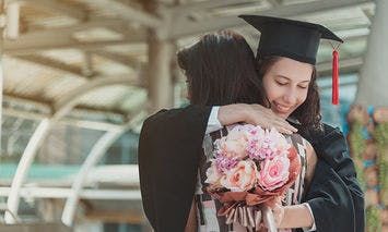 A graduate in a cap and gown hugs someone while holding a bouquet of pink flowers in an urban outdoor setting with a glass and steel structure in the background.